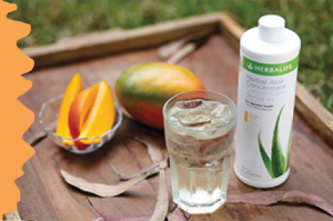 Un bicchiere di bevanda ghiacciata accanto a un mango e una bottiglia di aloe concentrato su un vassoio di legno.